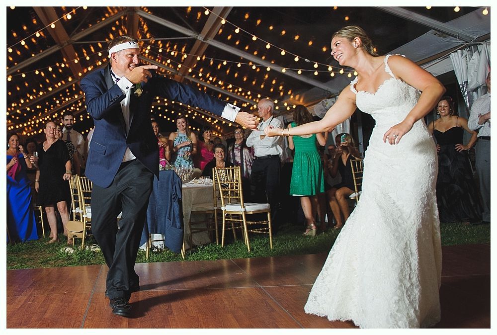 Bride and groom dancing at a wedding reception under string lights. Groom points; bride smiles in white gown.