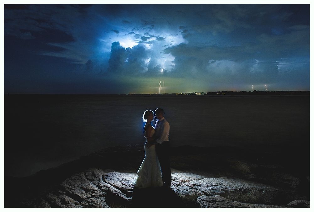 Couple embraces on a rocky shore at night, silhouetted against a dramatic lightning-filled sky.
