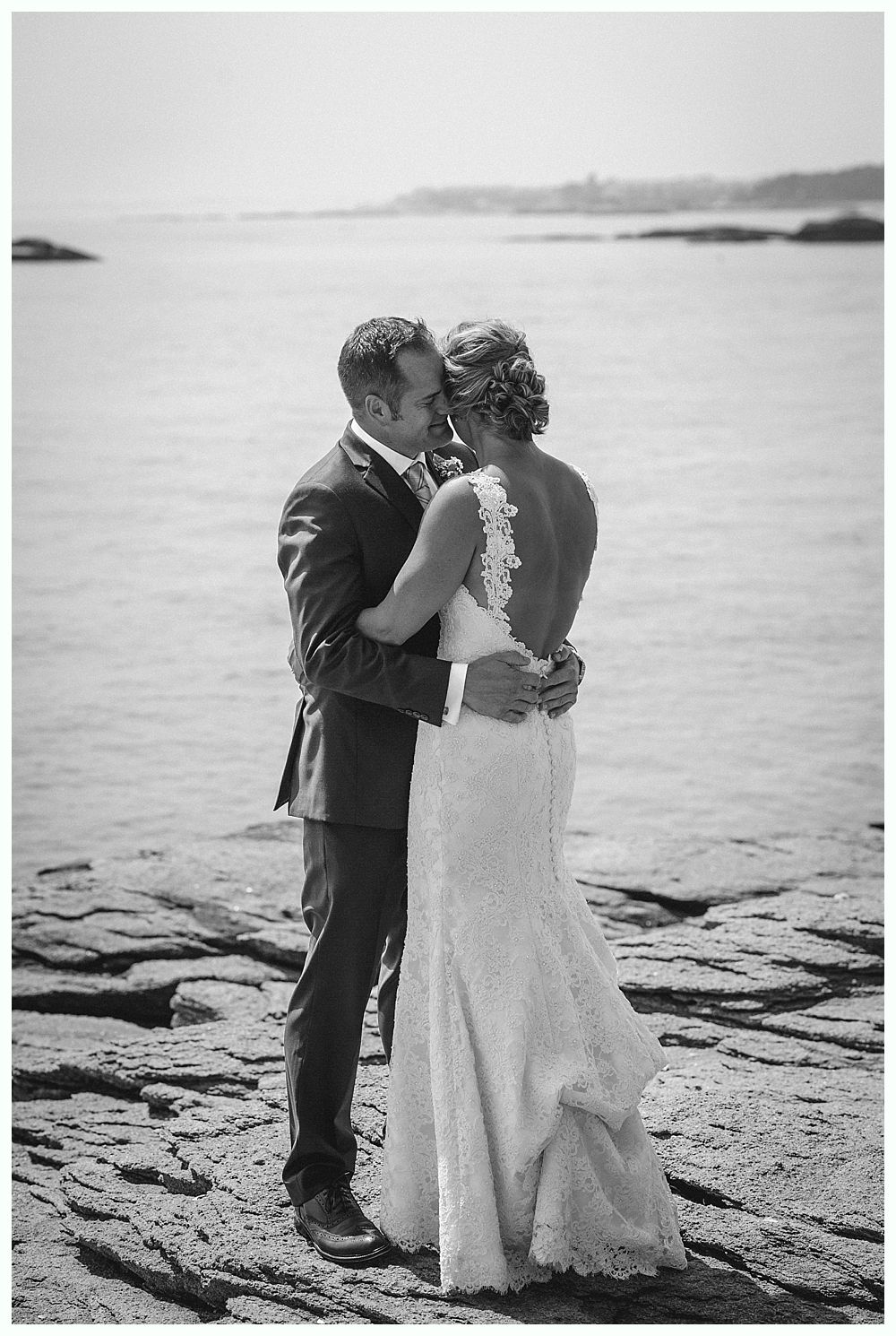 Bride and groom embrace on rocky shoreline near water, groom in suit, bride in lace dress.
