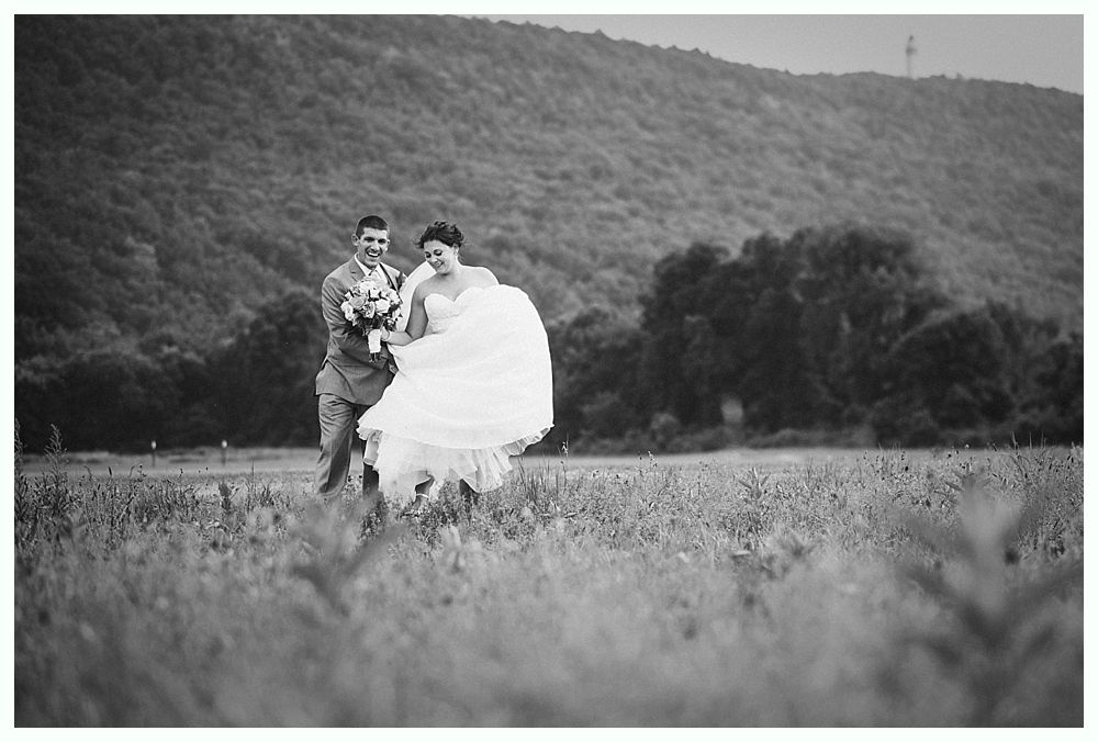 Bride and groom in wedding attire walking in a field, mountain backdrop. Black and white.