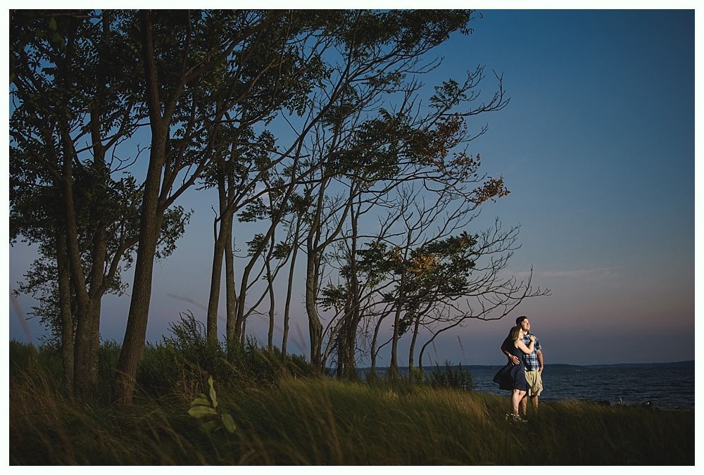 Couple standing on a grassy hill overlooking the ocean at dusk. Trees frame the view with a blue and orange sky.