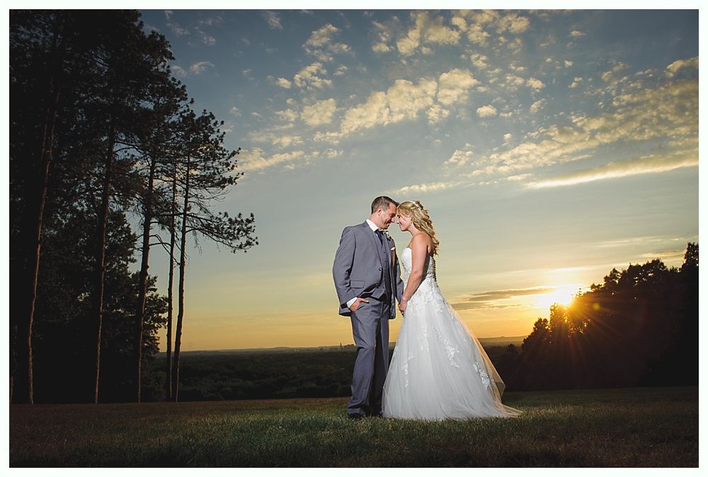 Couple embraces at sunset in a field; man in gray suit, woman in white dress.
