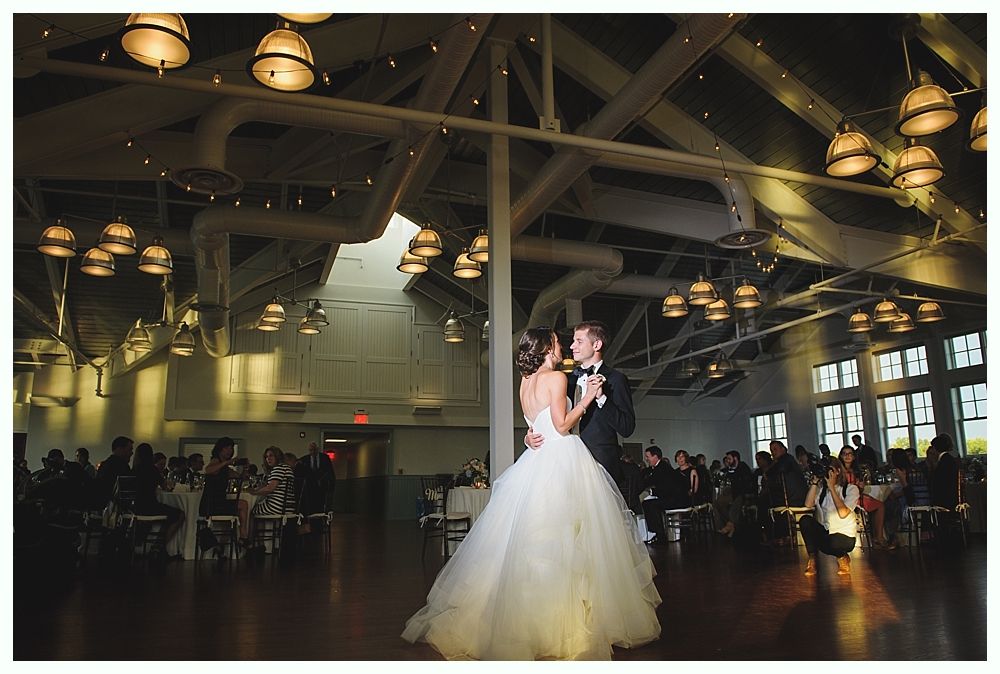 Bride and groom dance at a wedding reception. Venue lit with hanging lights; guests seated at tables.