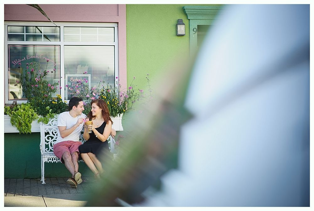 Couple sitting on a white bench, smiling and holding drinks outside a colorful building.