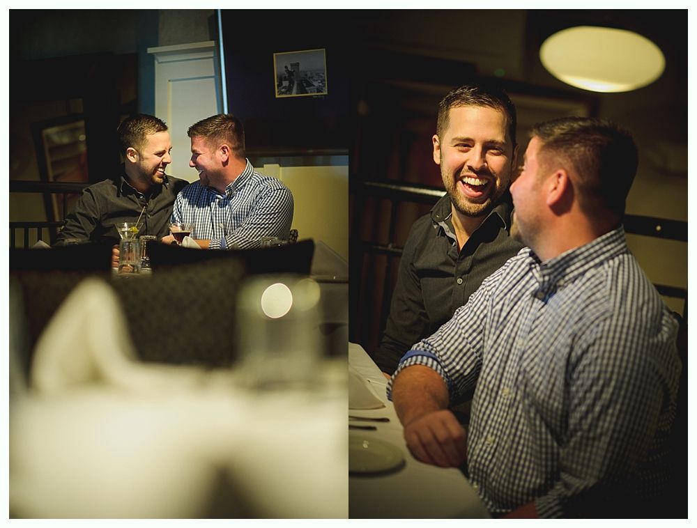 Two men laughing at a table in a restaurant. One wears a black shirt, the other a blue striped button-down.