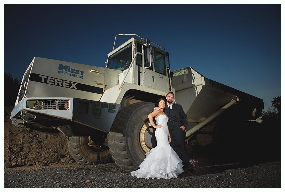 Couple in wedding attire posing beside large construction truck.