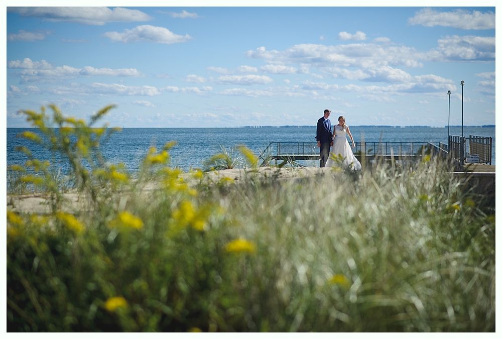 Couple on a dock near the ocean on a sunny day with wildflowers in the foreground.