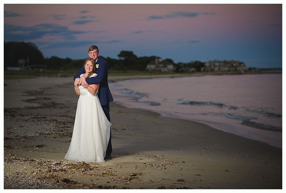Bride and groom embrace on a beach at dusk, ocean in background.