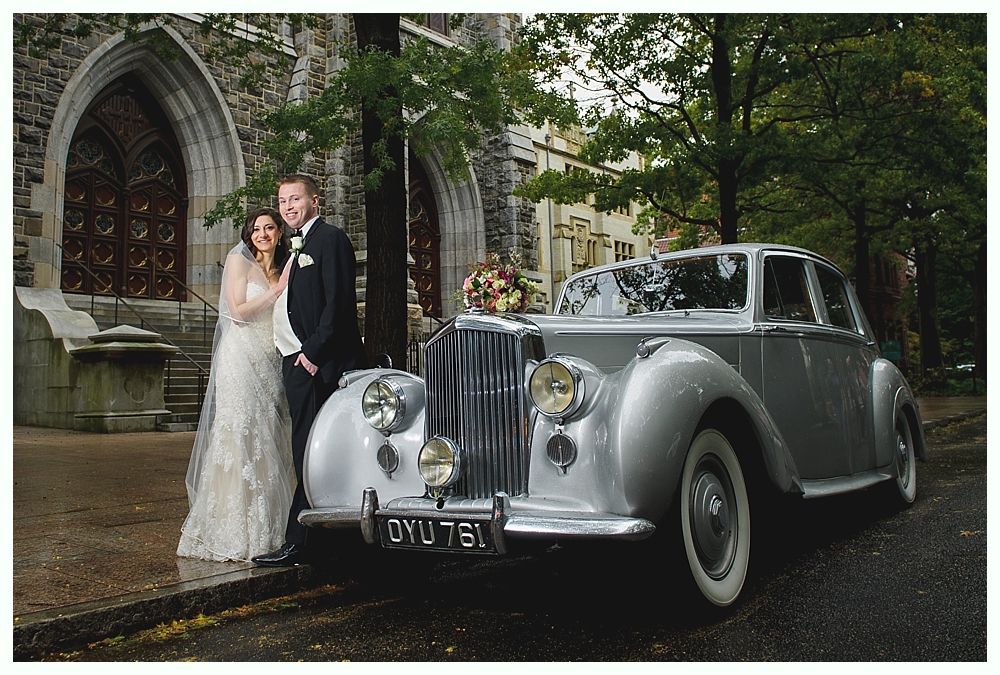 Newlyweds pose by a silver vintage car in front of a stone church. Bride smiles, groom looks ahead.