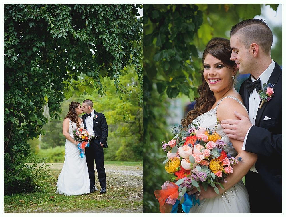 Bride and groom pose outdoors. Bride in white dress, holding bouquet. Groom in tuxedo, smiling. Green foliage background.