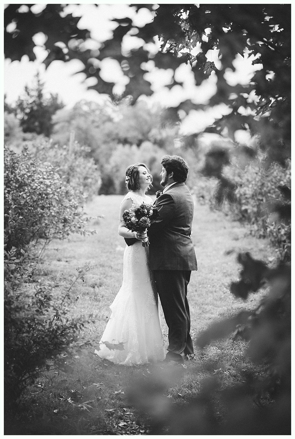 Bride and groom embrace in a field, trees framing the shot, soft lighting, black and white.