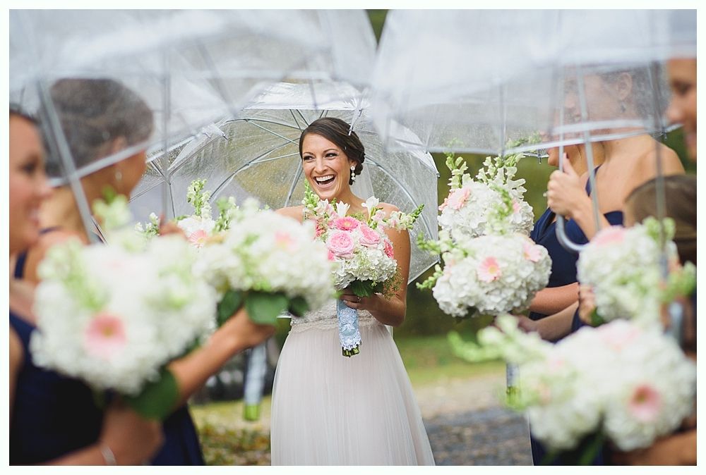 Bride laughs outdoors, surrounded by bridesmaids with bouquets and clear umbrellas.