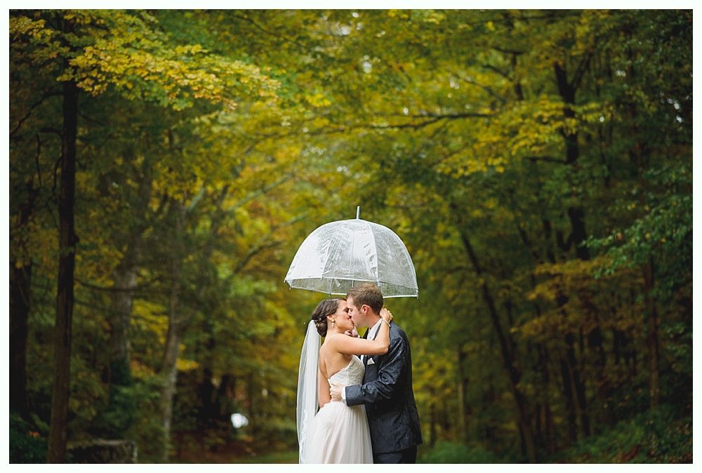 Newlyweds kiss under a clear umbrella in a wooded area; trees with fall foliage in the background.