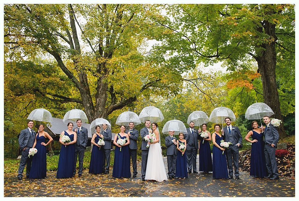 Wedding party poses outdoors under umbrellas; autumn foliage in background.