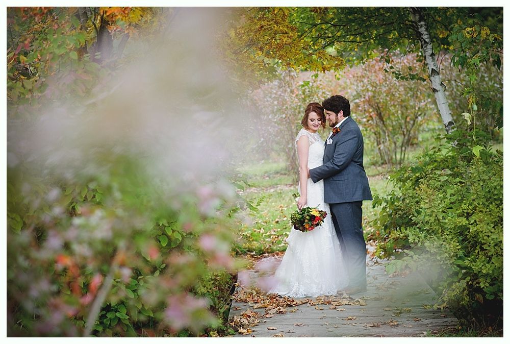 Couple embracing on a wooden path surrounded by autumn foliage; bride in white gown, groom in suit.