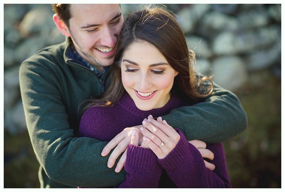 Man hugging woman; both smiling, in a natural setting. He wears green, she wears purple; engagement ring visible.