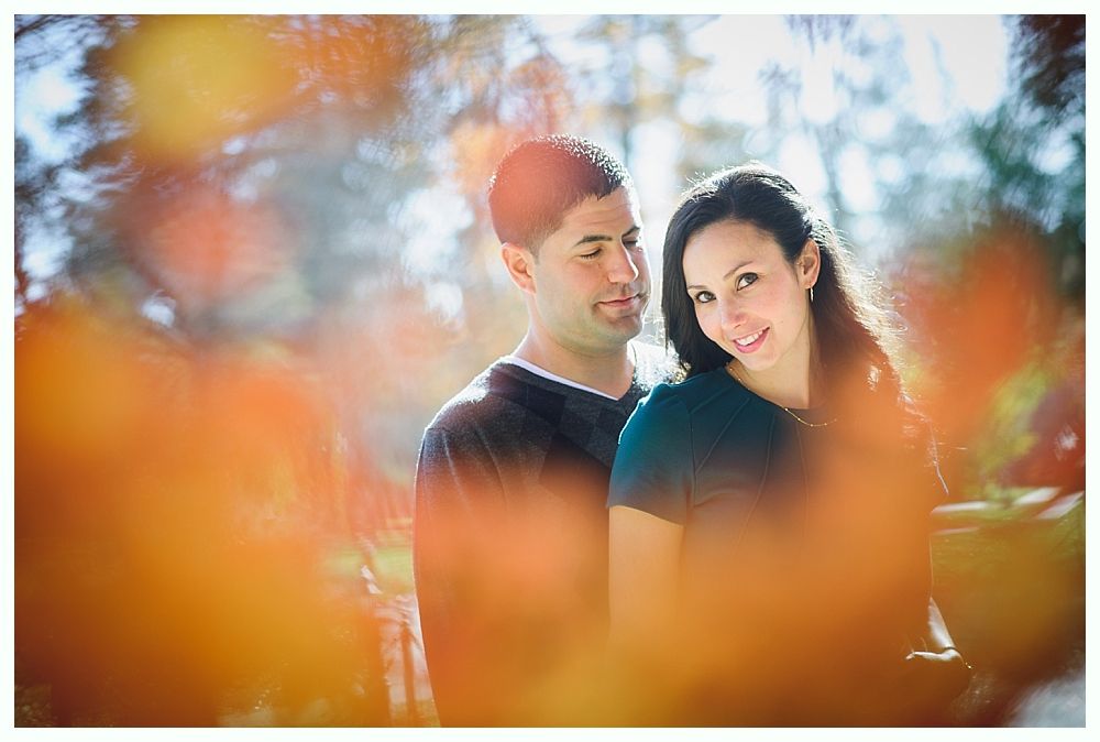 Couple smiles in a park. Woman in teal shirt looks at the camera, man looks at her. Fall foliage in foreground.