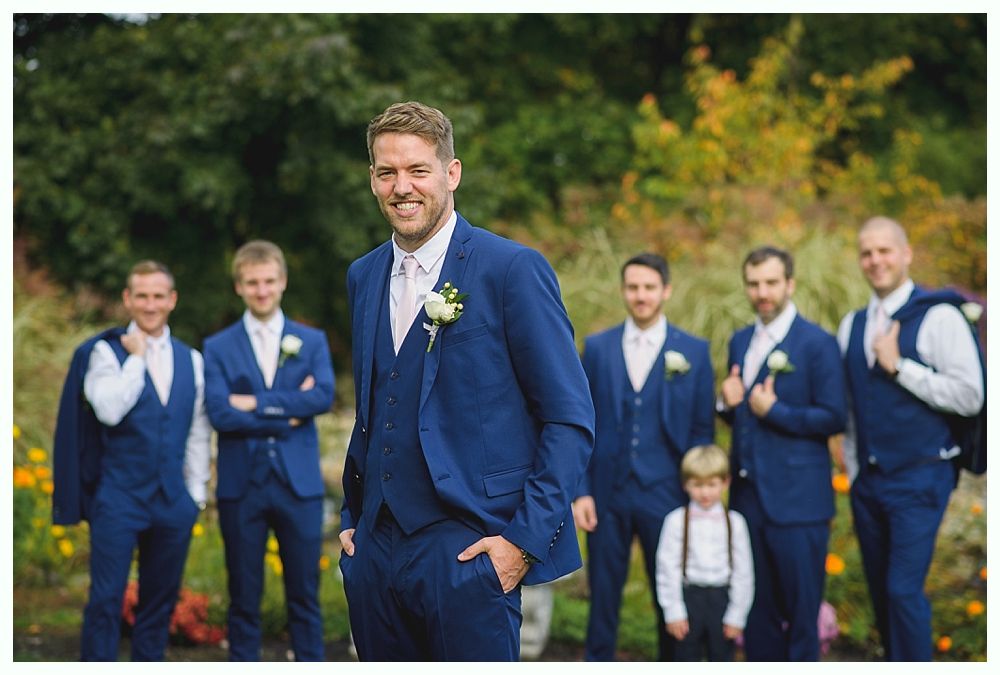 Groom and groomsmen in blue suits pose outdoors, smiling. The groom has hand in pocket, a light pink tie, and a boutonniere.