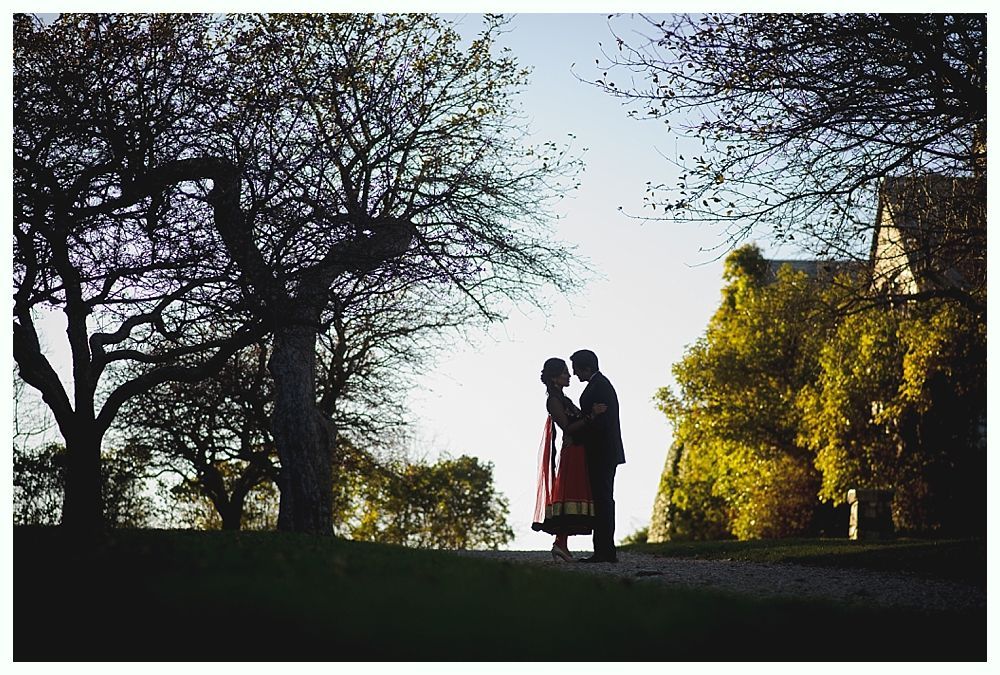 Couple embraces on a path, silhouetted against a bright sky, framed by trees and a building.