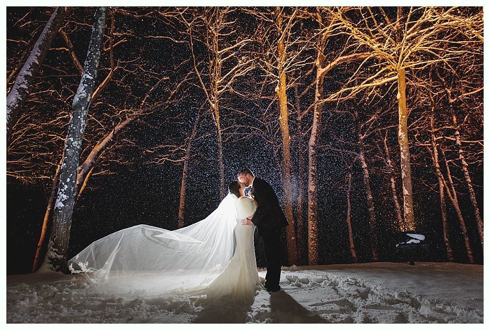 Couple kissing in snow-covered forest at night, bride's veil flowing, trees lit with golden lights.