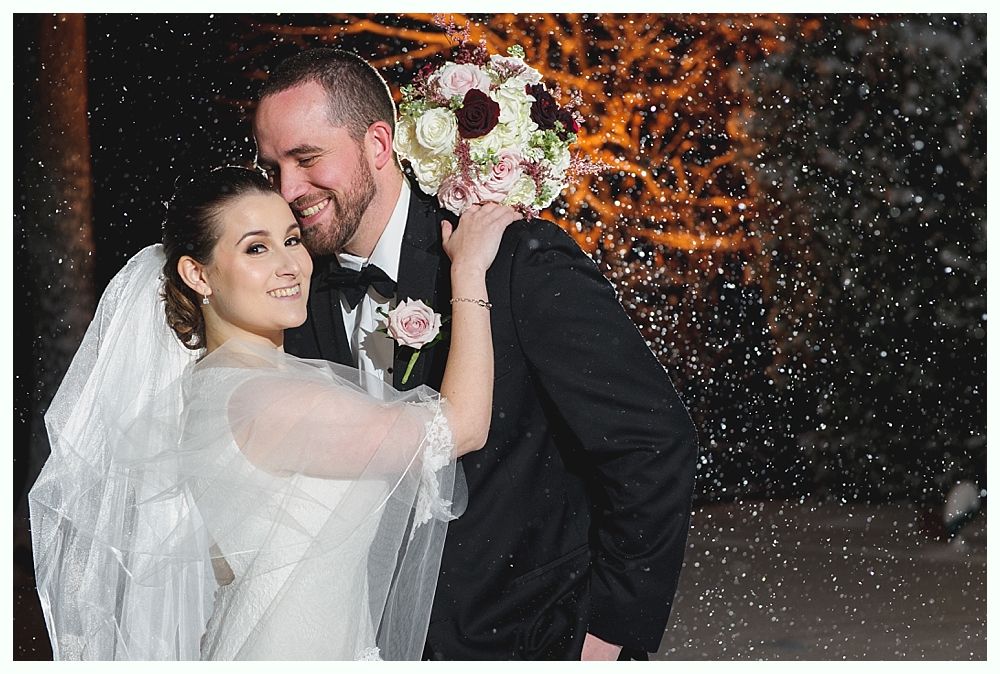 Bride and groom embrace in snow. Bride in white dress and veil, groom in black suit, both smiling.