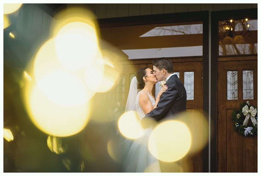 Couple kissing in front of a wooden door, blurred yellow lights in the foreground.
