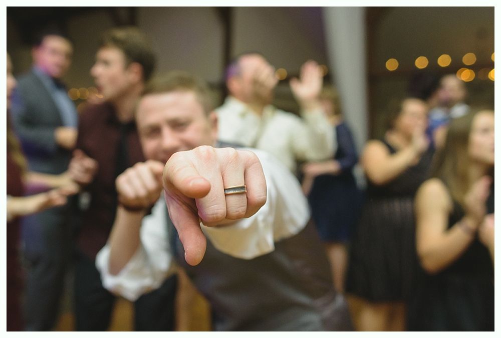 Man points finger towards camera at a party, wearing a vest and ring, other people dancing in the background.