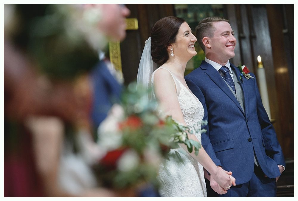 Bride and groom holding hands, smiling during their wedding ceremony. They are inside a church.