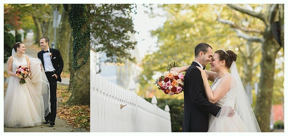 Wedding couple in formal attire, walking and embracing near white fence and trees.