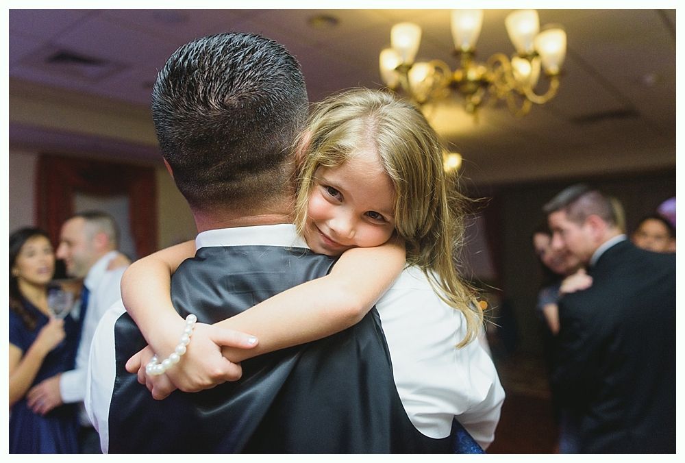Girl hugs a man, resting her head on his shoulder, smiling. Ballroom with people dancing in background.
