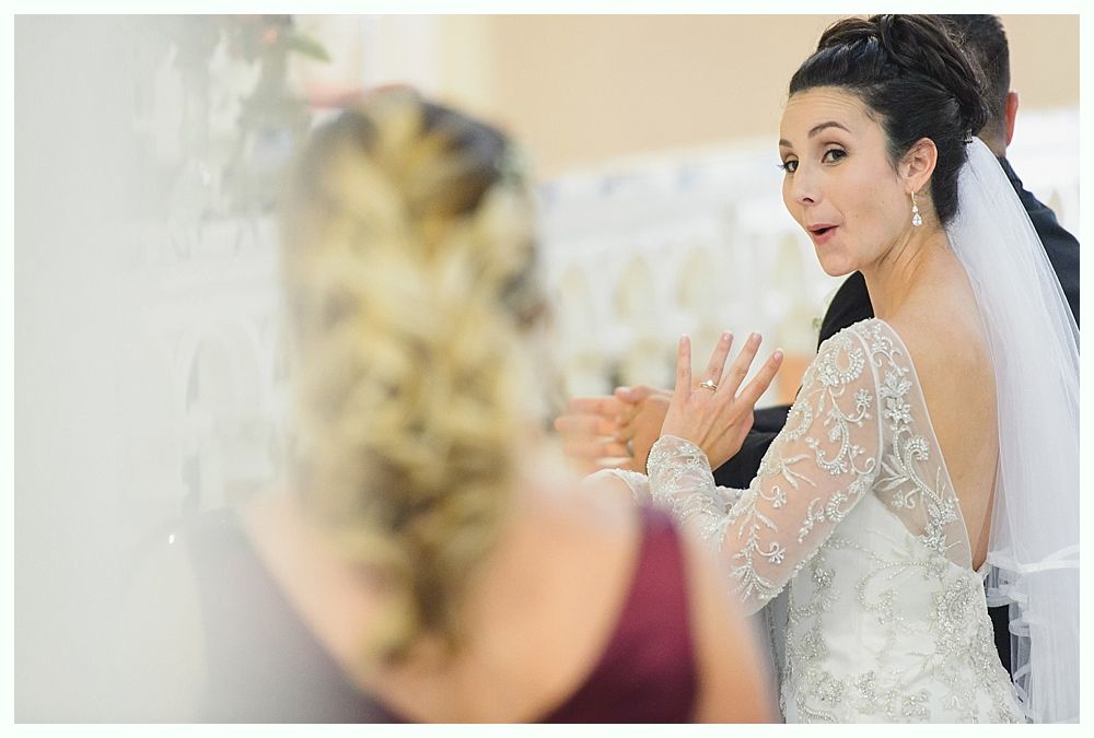 Bride in wedding dress, looking surprised, raising hand. Veil, long-sleeved dress, and a person in a burgundy top visible.