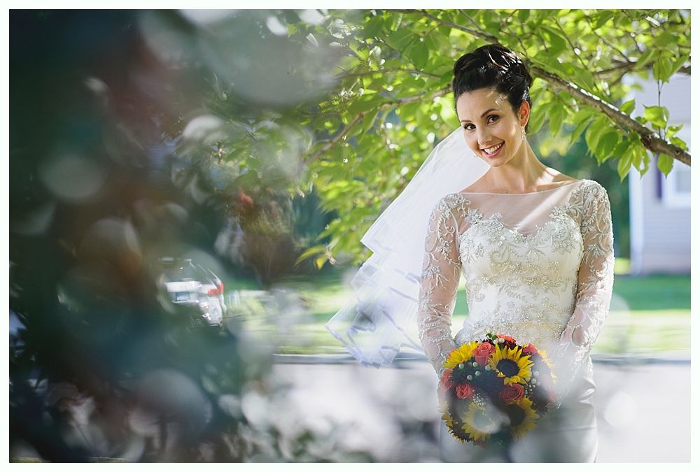 Bride in white dress holding bouquet, smiling in front of green trees with sun flares.