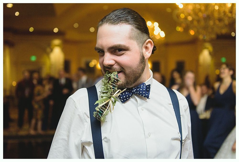 Man in white shirt, blue polka dot bow tie, suspenders, holding greenery in mouth, at event.