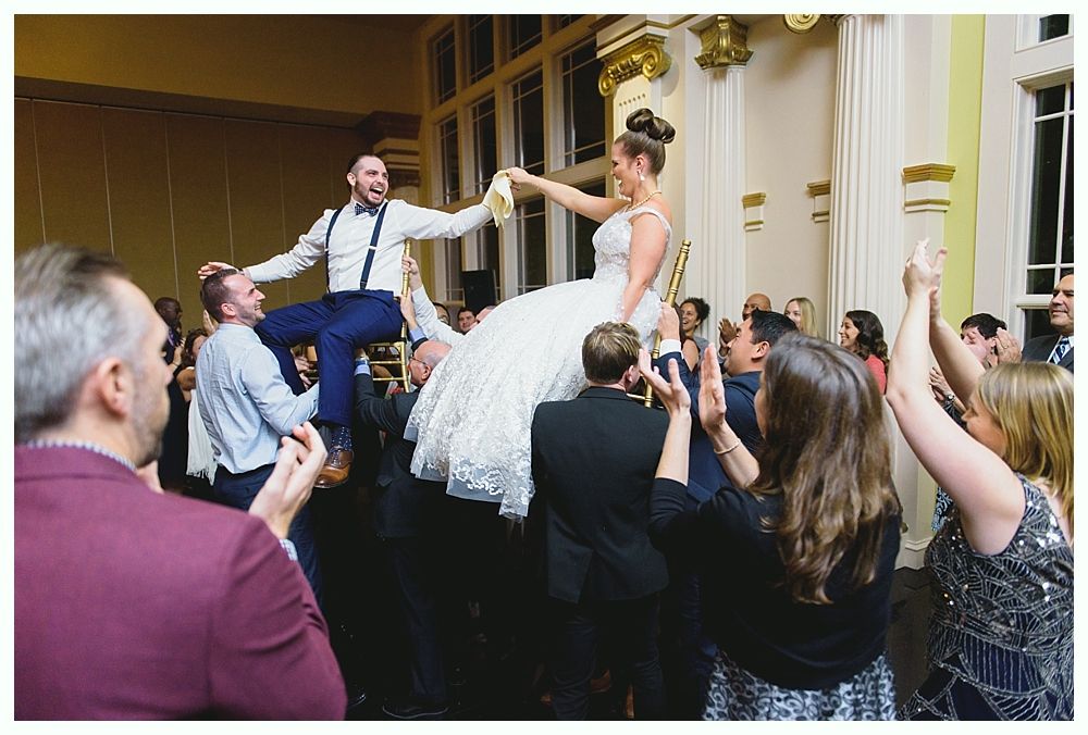 Wedding guests lift the bride and groom on chairs during a celebration. Bright room with large windows.