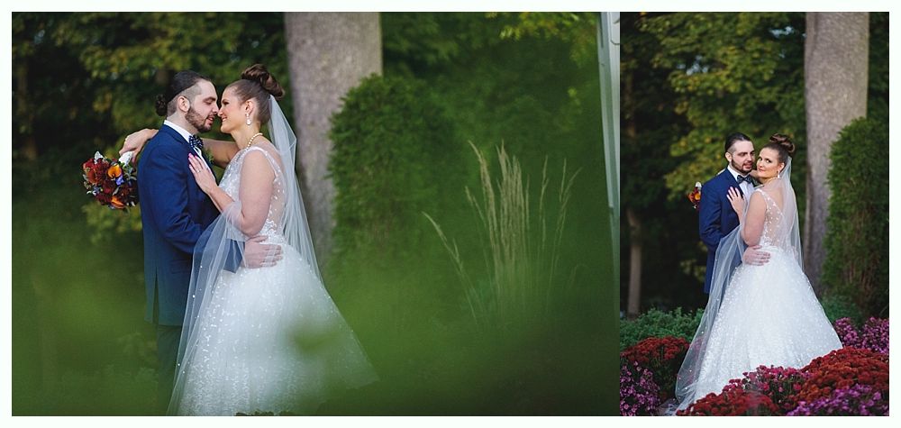 Bride and groom embrace outdoors, she in a white dress and veil, he in a blue suit. Green foliage frames the couple.
