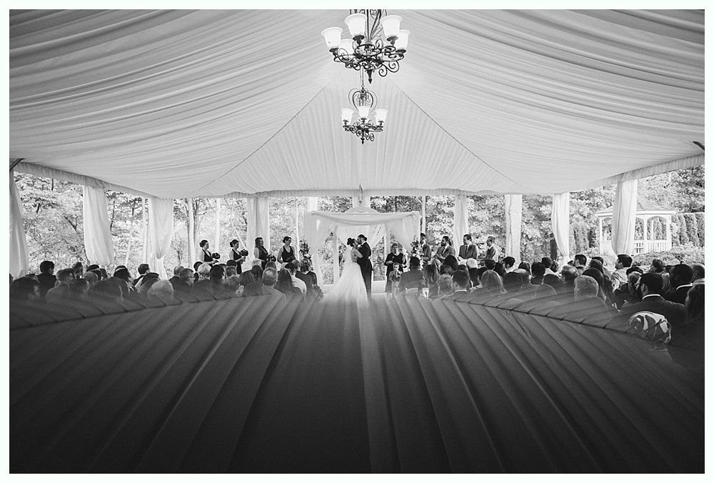 Black and white photo of a wedding ceremony under a tent. A couple stands center, facing an audience.