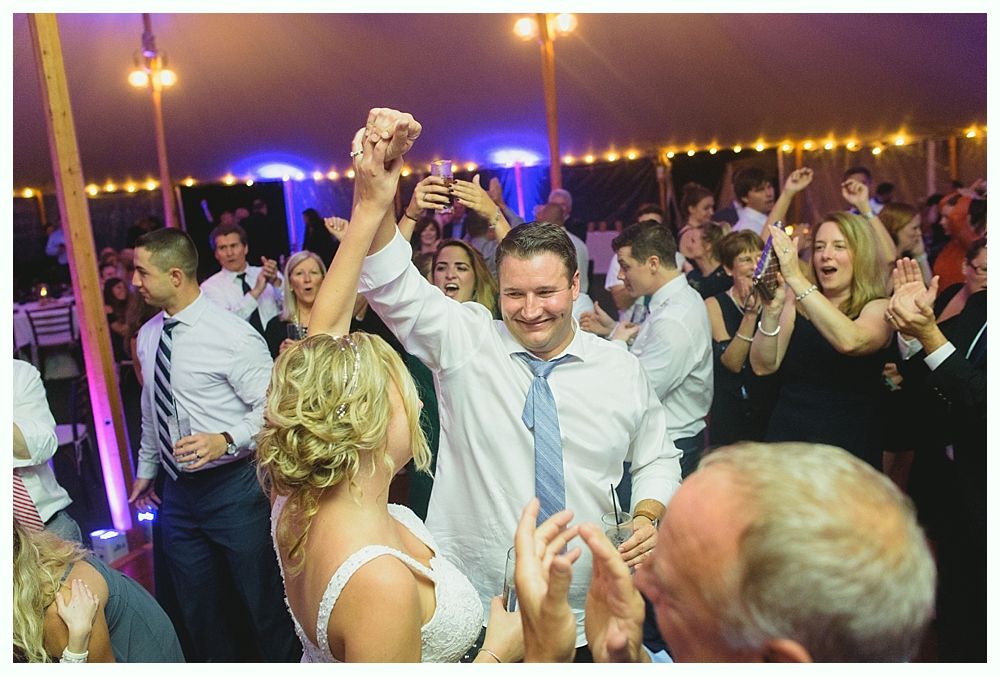 People celebrating at a wedding reception, some dancing and cheering. Lit tent with string lights.