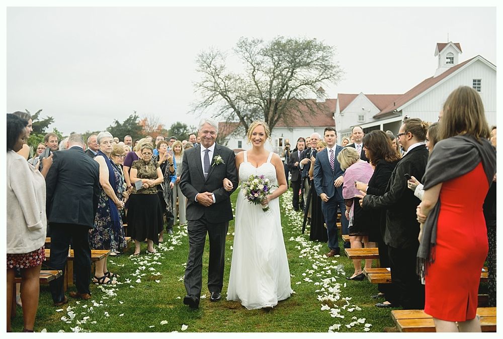 Bride and father walk down aisle, lined with guests and rose petals, towards white building under cloudy sky.