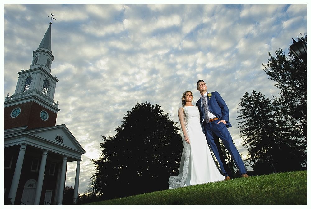 Bride and groom pose on grassy hill with church in the background, cloudy sky overhead.