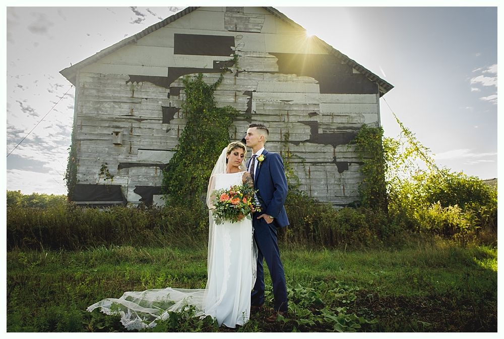 Newlyweds pose in front of a weathered barn on a sunny day. Bride in white dress, groom in blue suit.