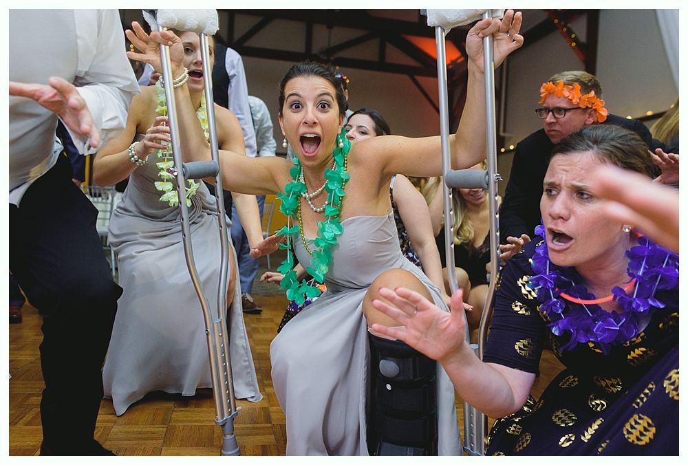 Woman in formal dress with leg brace on crutches dancing, surrounded by others at a party; all are excited.