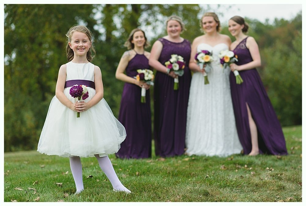Flower girl in white dress and purple sash, holding flowers, smiles at the camera, with bridesmaids and bride in background.