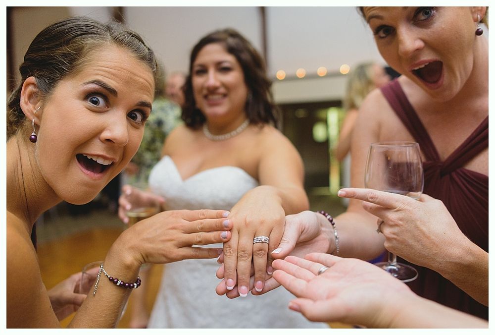 Three women at a wedding excitedly look at a ring on a bride's hand. One points, all have surprised expressions.
