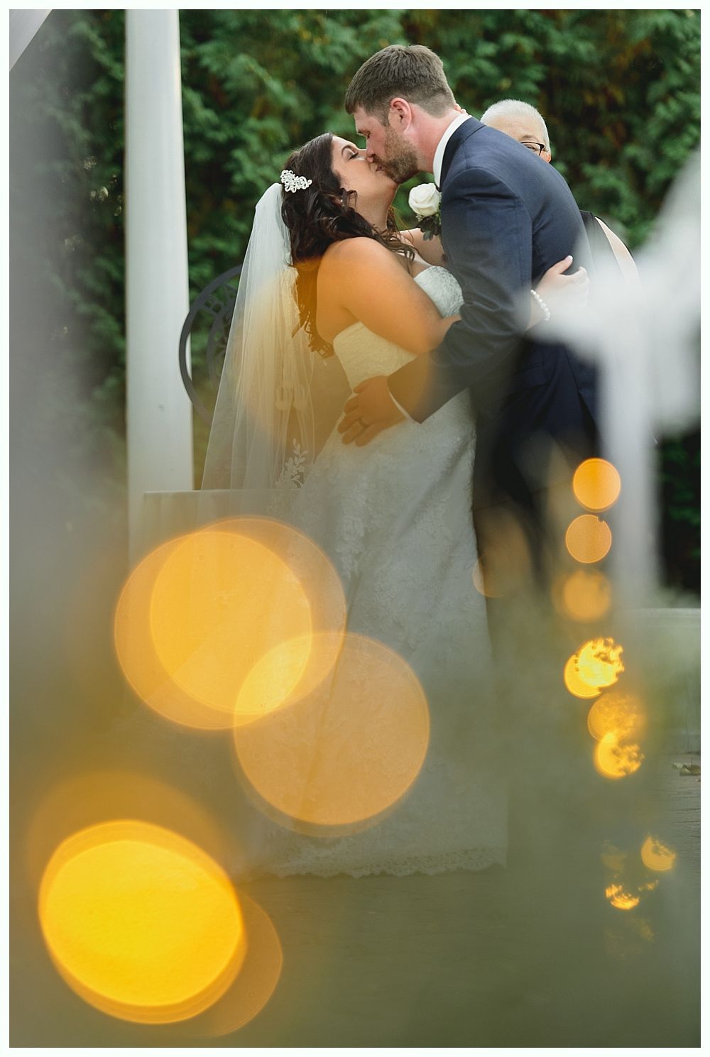 Bride and groom kissing during wedding ceremony near a white pillar; bokeh lights in foreground.