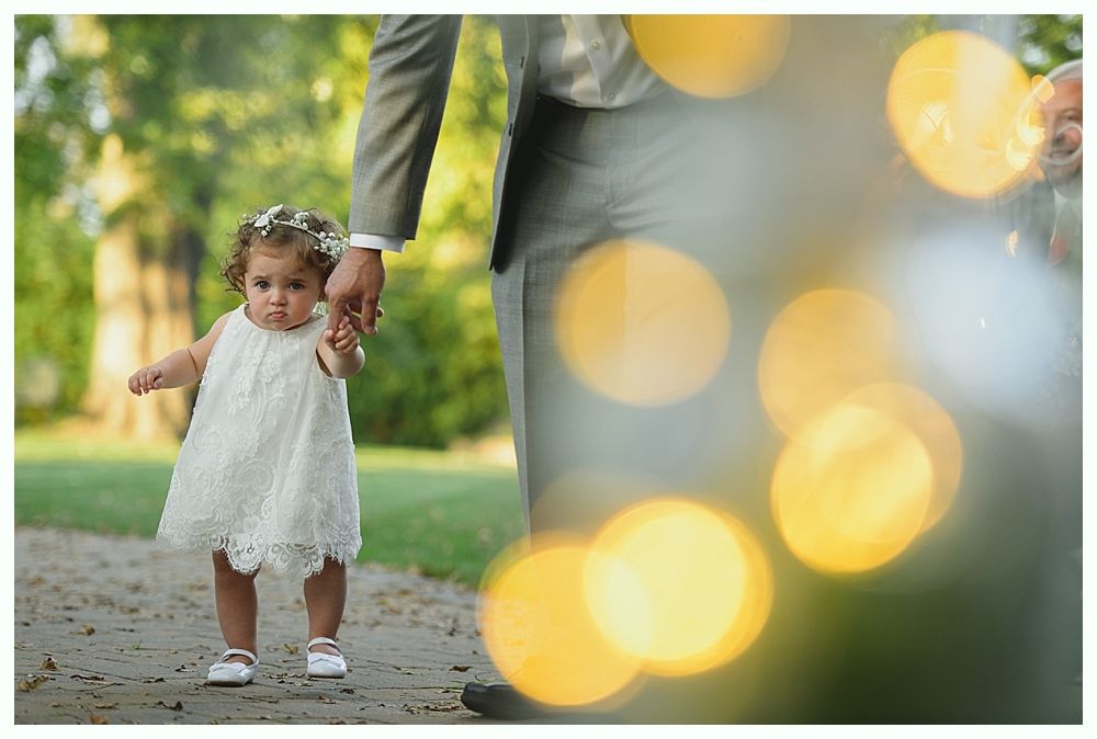 Young girl in white dress holds hands with a person in a suit, walking outside with soft bokeh lights.