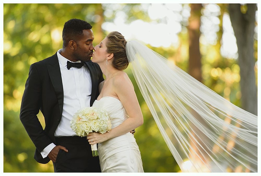 Couple kissing; the bride in white dress, veil, holding flowers, groom in black tuxedo, outdoor setting.