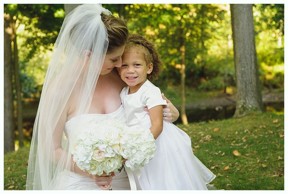 Bride in white dress holding flower bouquet with flower girl, outdoors in front of trees.