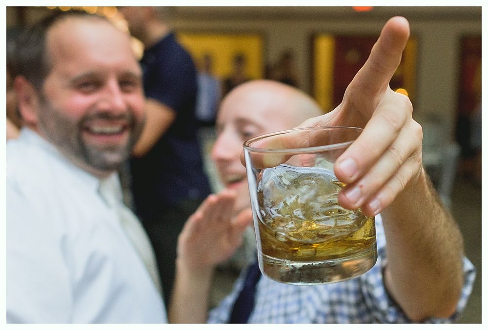 Man holding up a whiskey on the rocks, smiling with another man in background.