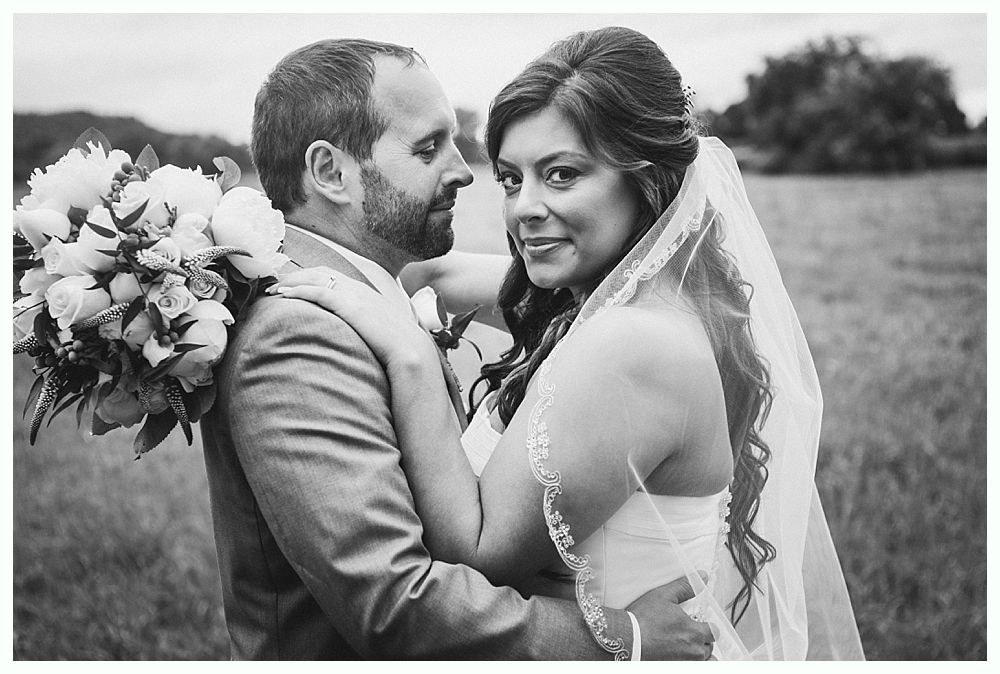 Bride and groom embrace in a field, woman smiling, holding bouquet, man in suit.