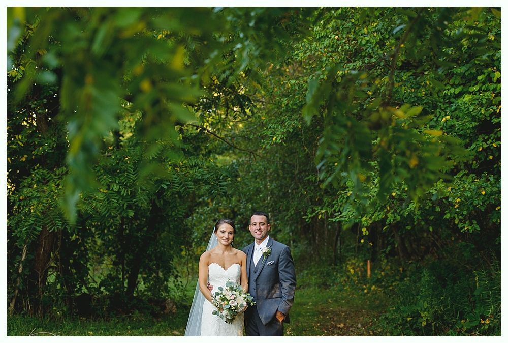 Bride and groom pose in a lush, green forest. The bride wears a white dress and veil, and the groom a gray suit.